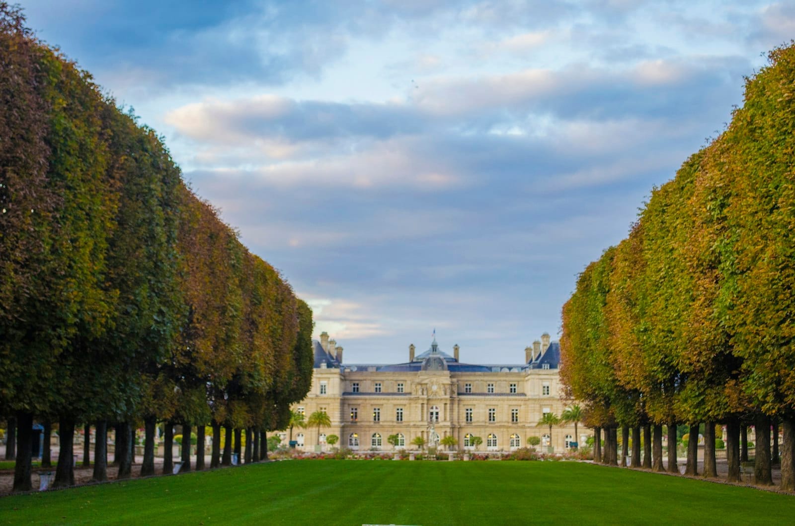 Elegante Baumreihen mit Herbstblättern rahmen den Rasen ein, der unter einem teilweise bewölkten Himmel zum Palais du Luxembourg in Paris führt.