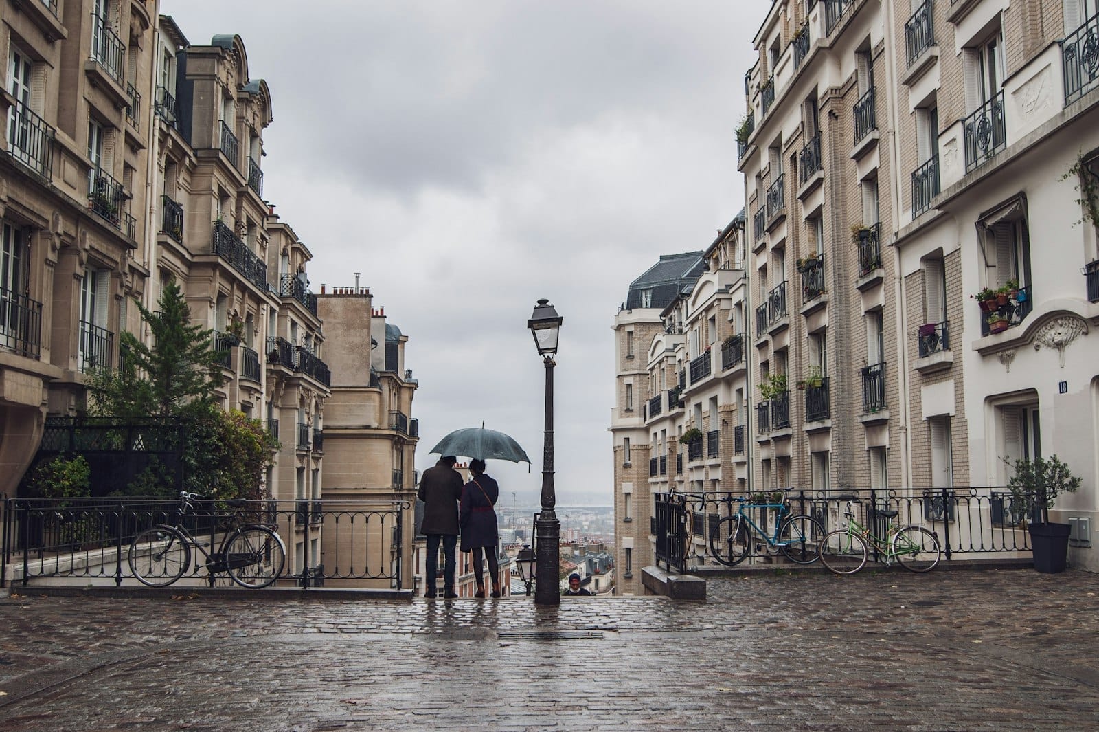 Zwei Personen unter einem Regenschirm stehen auf einer regnerischen Pariser Straße, gesäumt von klassischen Apartmentgebäuden und nassen Kopfsteinpflastersteinen.