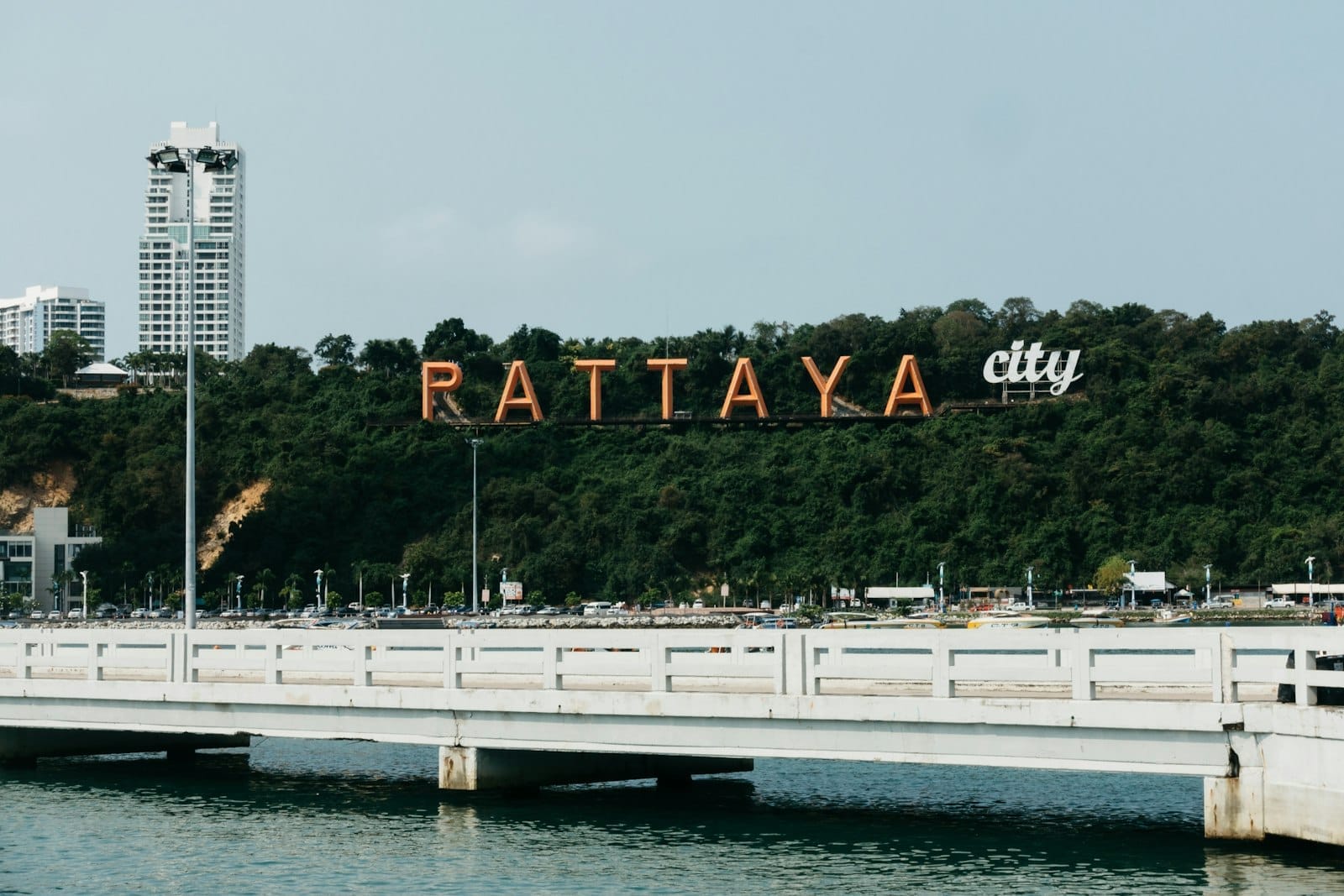 Grande letreiro laranja de Pattaya City em uma colina verde, edifícios altos brancos ao fundo, com uma ponte cruzando águas calmas em primeiro plano.