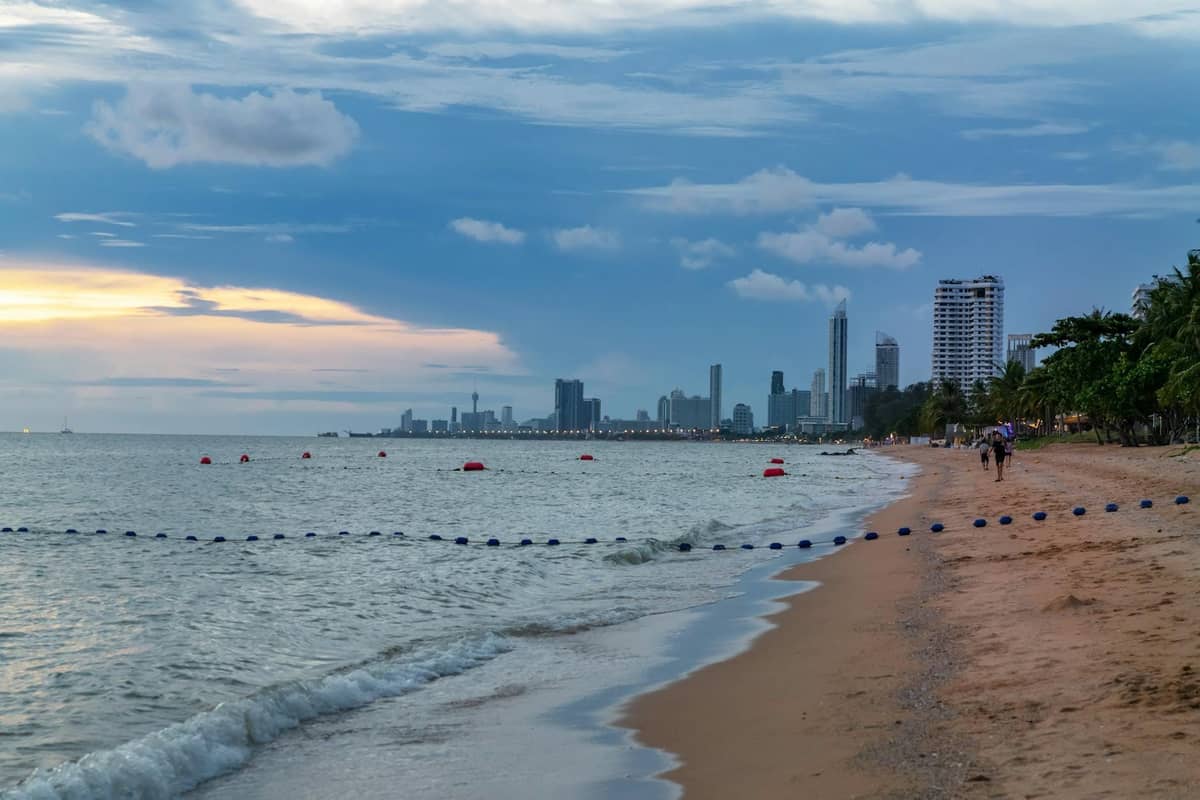 Pattaya beachfront with sandy shore, city skyline in the distance, ocean waves, and a partly cloudy sky, showing typical coastal weather.