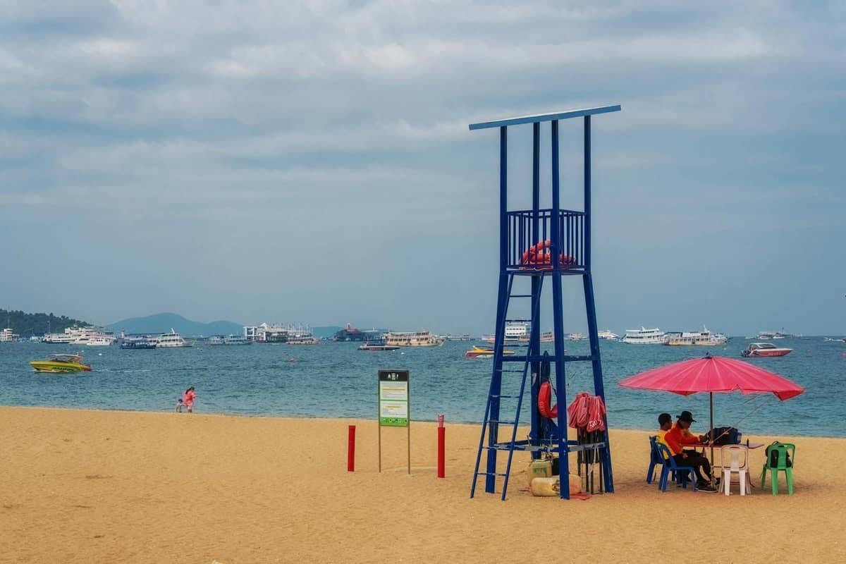 Sandy Pattaya beach with a blue lifeguard tower, pink umbrella, and cloudy skies, with boats anchored in the background.