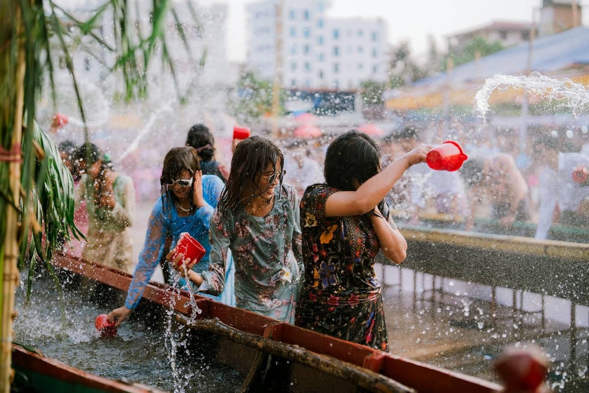 Women playing with water and splashing each other at an outdoor Songkran festival event in Pattaya, with water containers and city buildings visible.