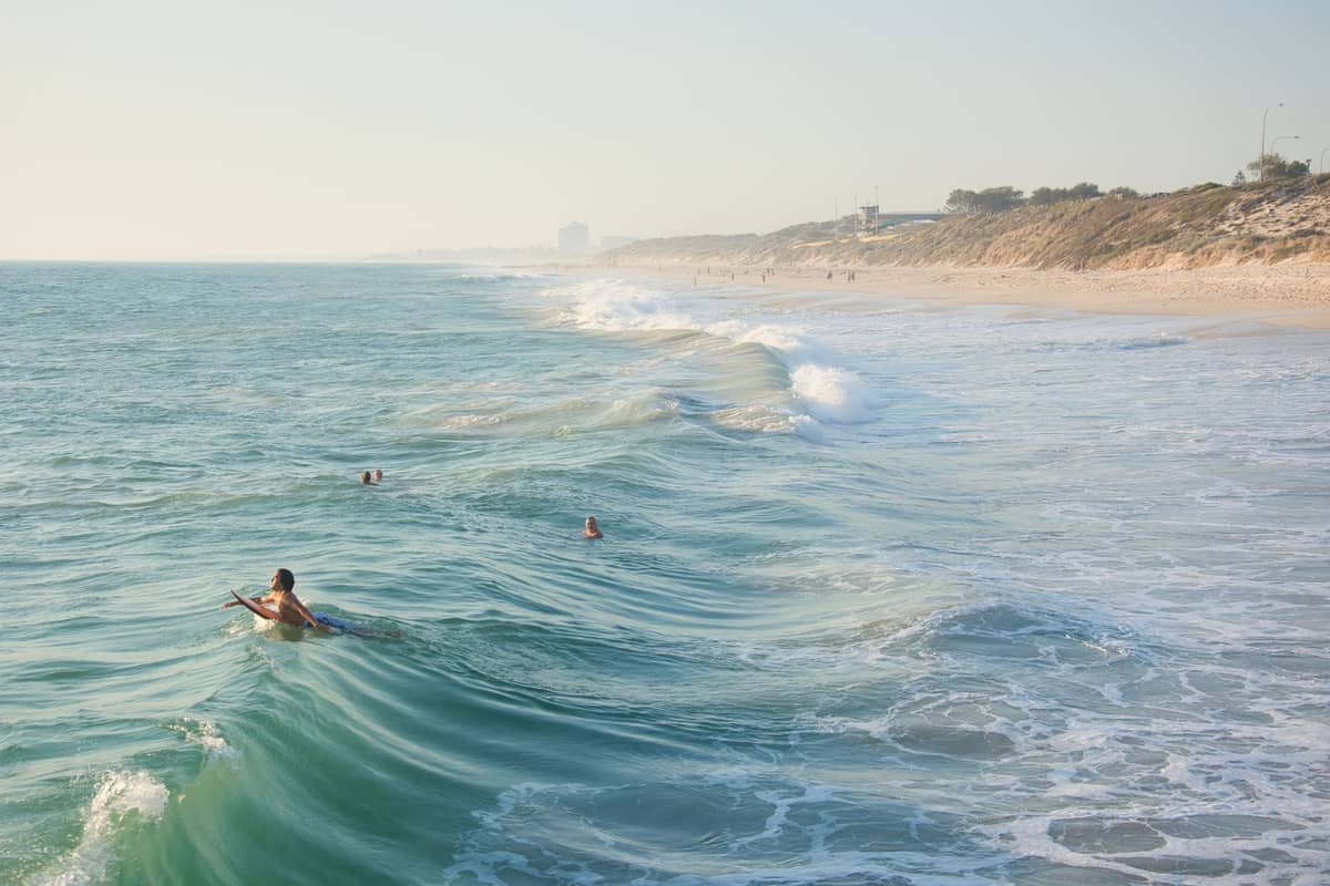 Diversi nuotatori e una persona su una tavola da surf tra le onde gentili di una spiaggia sabbiosa di Perth, con dune di sabbia sullo sfondo.