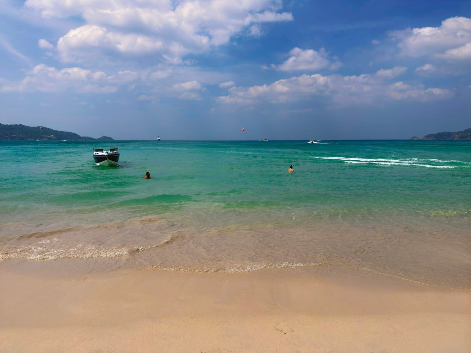 Clear turquoise water with a boat and a few swimmers, sandy beach, and blue sky with scattered clouds in Phuket.