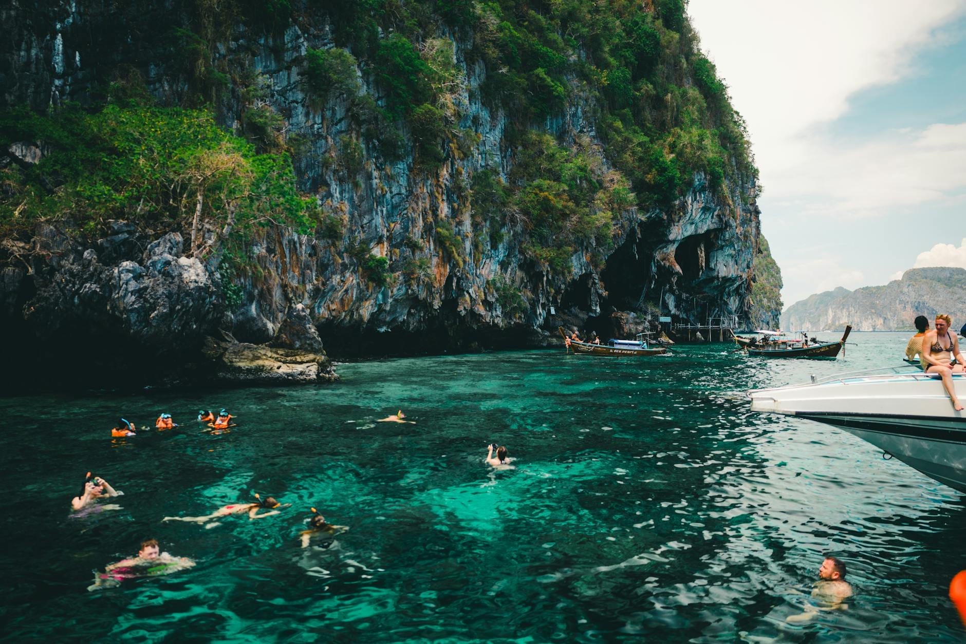 Tourists swimming and snorkeling in clear turquoise water near dramatic limestone cliffs, with boats and lush greenery in the background, typical of Phuket’s adventure activities.