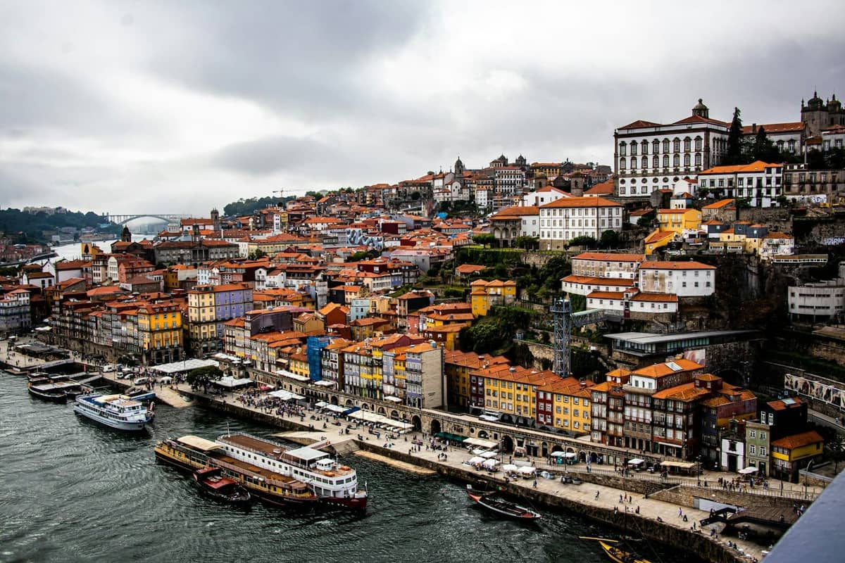 Vivace lungofiume di Porto con edifici storici colorati, barche e folle lungo il waterfront in una giornata nuvolosa.