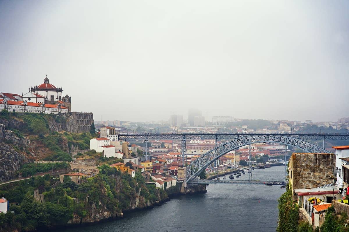 La nebbia avvolge lo skyline di Porto, con il fiume Douro, l'iconica Ponte Dom Luís I e gli edifici sulle colline visibili sotto un cielo nuvoloso.