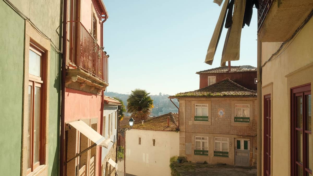 Una tranquilla strada soleggiata di Porto fiancheggiata da case colorate e balconi, panni stesi fuori e un accenno del fiume Douro in lontananza.