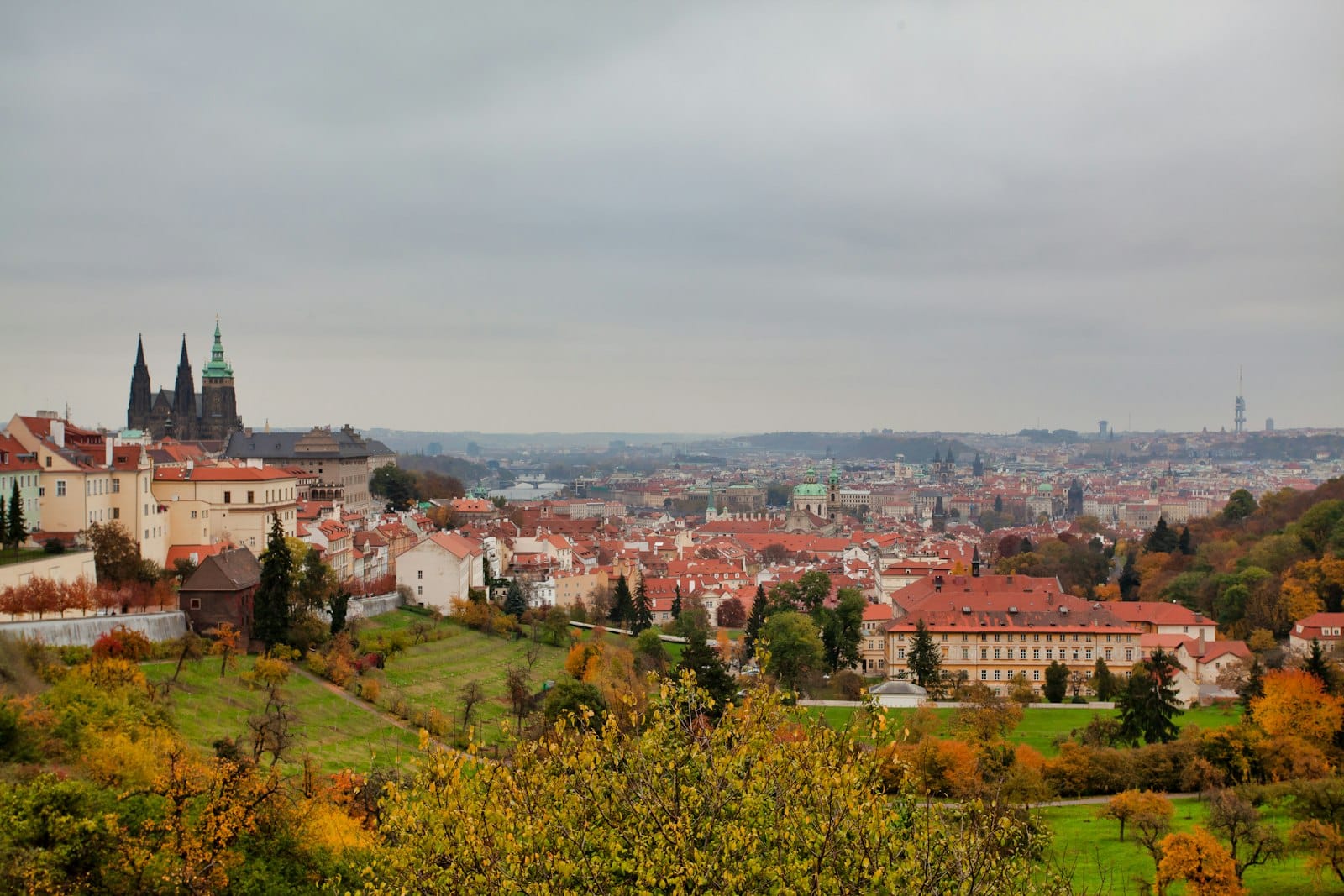 Bewölkter Panoramablick auf Prag mit herbstlichem Laub, Stadtdächern und der Prager Burg links unter bedecktem Himmel.