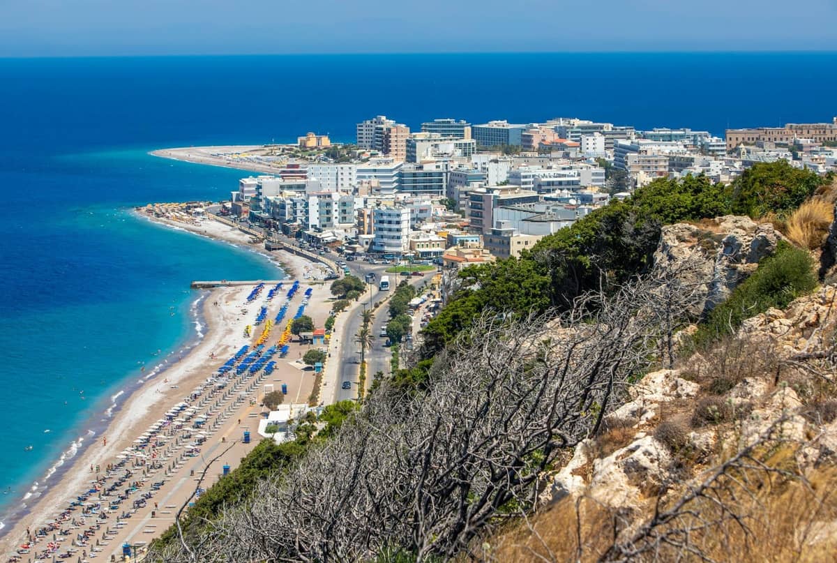 Aerial view of a busy summer beach with umbrellas and sunbeds in Rhodes, lined by coastal buildings and bright blue sea.