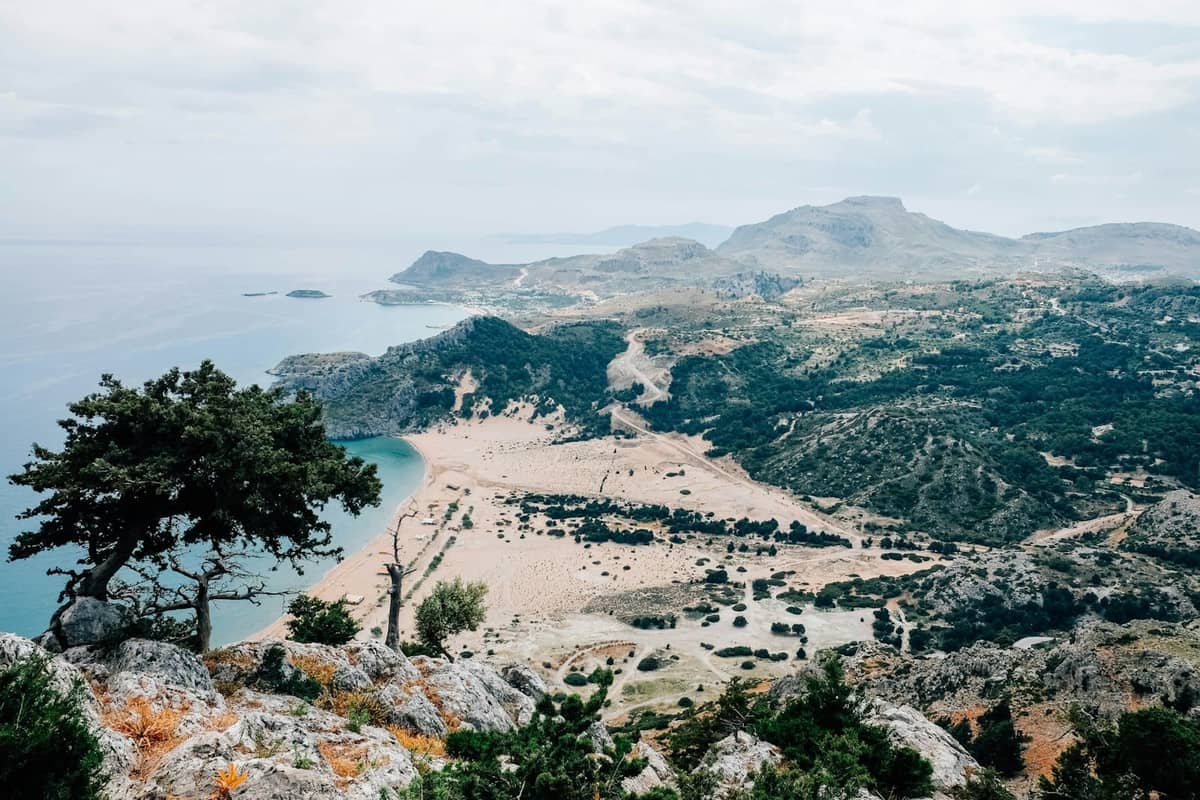 Panoramic view of a Rhodes coastline with sandy beaches, clear blue sea, and rugged mountains under a cloudy sky.