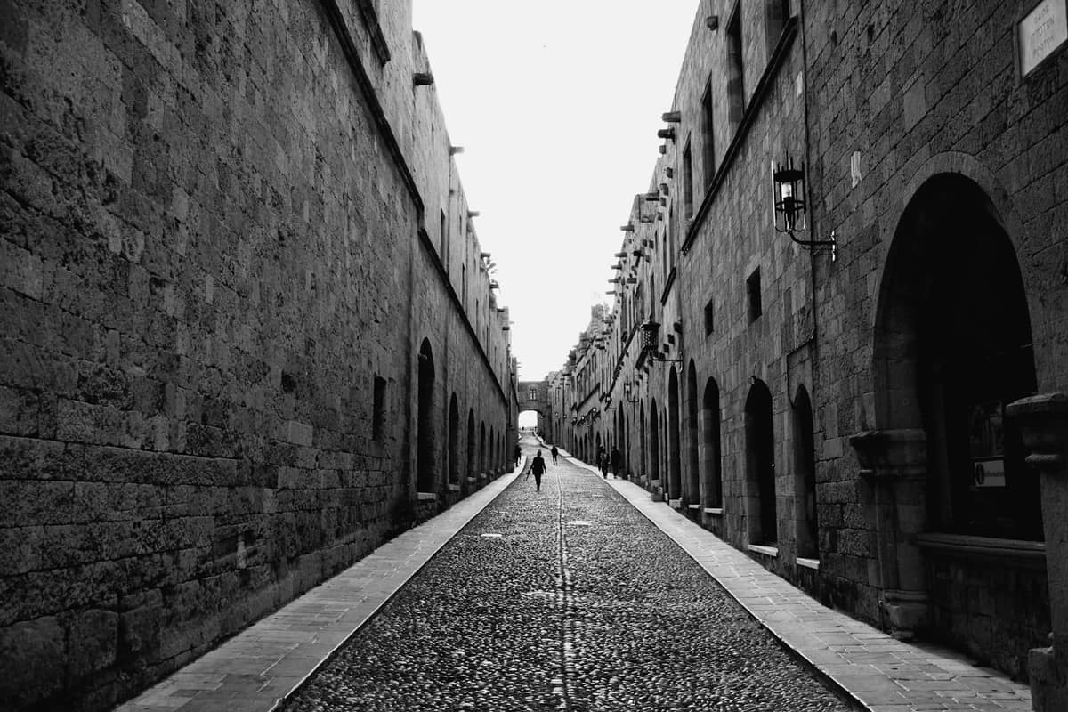 Wide cobbled medieval street in Rhodes lined with stone buildings under overcast winter sky, one person walking in the distance.
