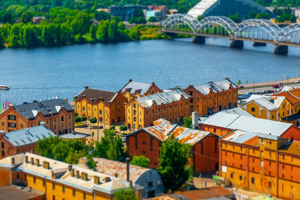 Luftaufnahme der alten Lagerhäuser Rigas am Fluss Daugava, mit einer historischen Bogeneisenbahnbrücke und üppigem Grün im Hintergrund.