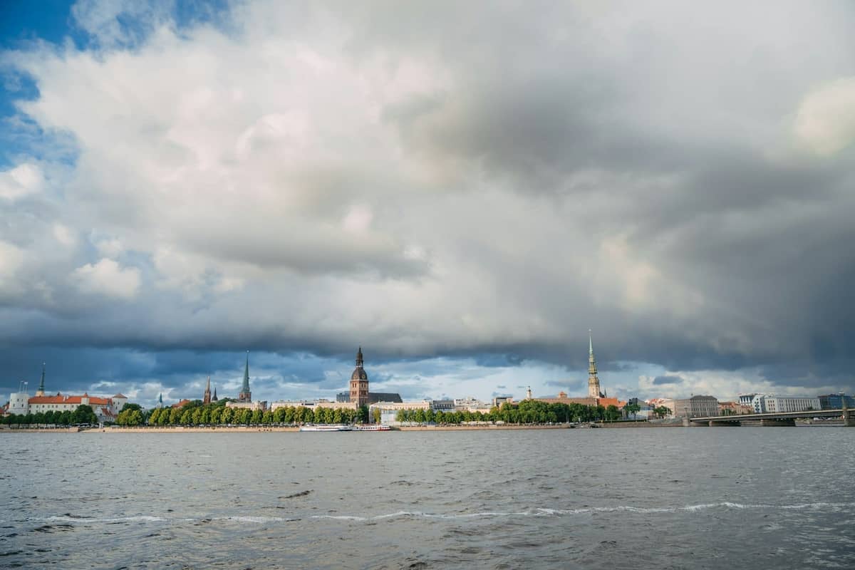 Weitwinkelansicht der Skyline von Riga über die Daugava mit sichtbarem Frühlingslaub und ikonischen Kirchturmspitzen unter dramatischen Wolken.