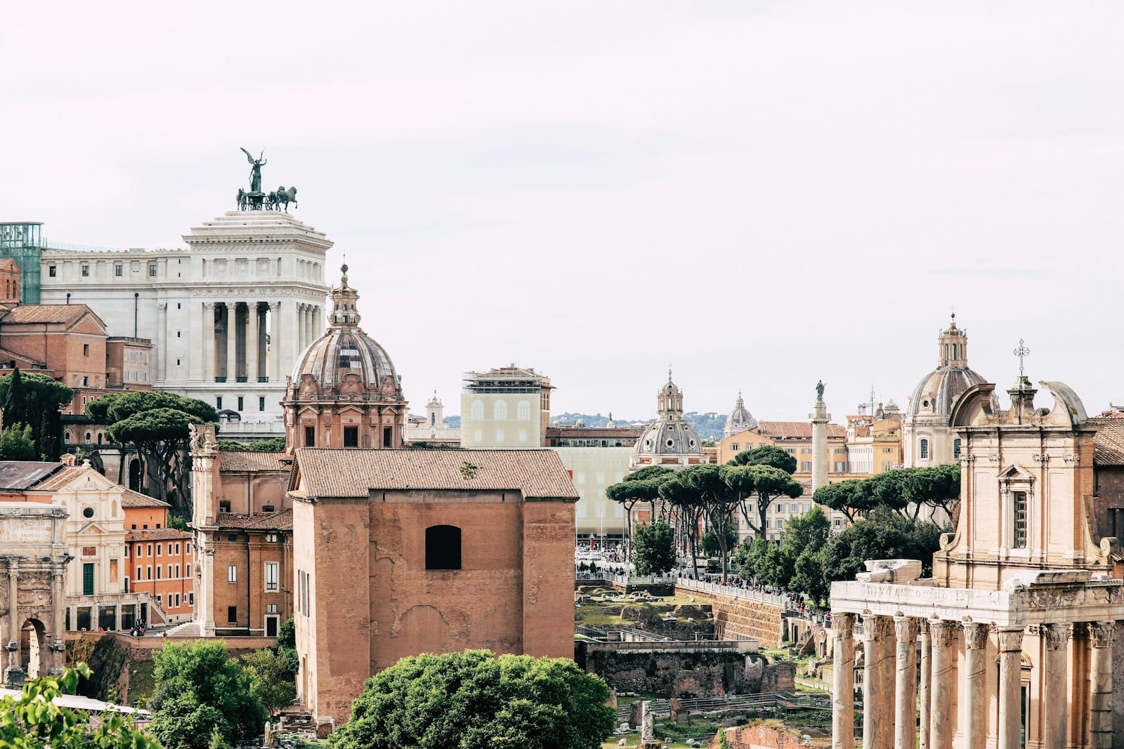 Panoramic view of Rome with historic domes, ancient ruins, and lush green trees on a bright, clear day.