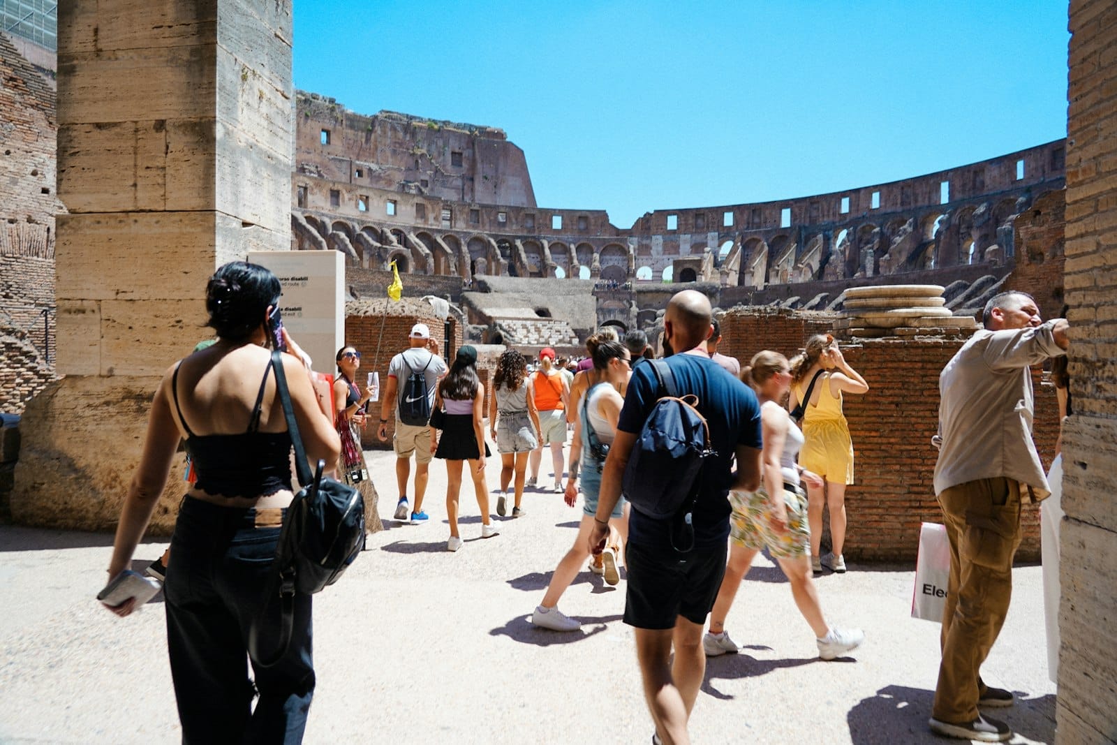 Tourists inside the sunlit Colosseum wearing summer clothes, with bright blue sky and ancient stone walls visible.