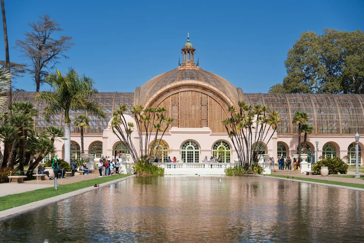 วิวของ Botanical Building และสระสะท้อนน้ำใน Balboa Park ภายใต้ท้องฟ้าสีฟ้าใส พร้อมต้นปาล์มและนักท่องเที่ยวที่เพลิดเพลินกับสภาพอากาศแดดจ้า