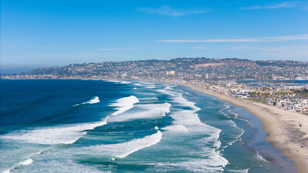 ภาพทางอากาศกว้างของแนวชายฝั่ง San Diego พร้อมหาดทรายกว้าง คลื่นมหาสมุทรสีฟ้า และย่านชายฝั่งที่คึกคักภายใต้ท้องฟ้าฤดูร้อนที่แจ่มใส