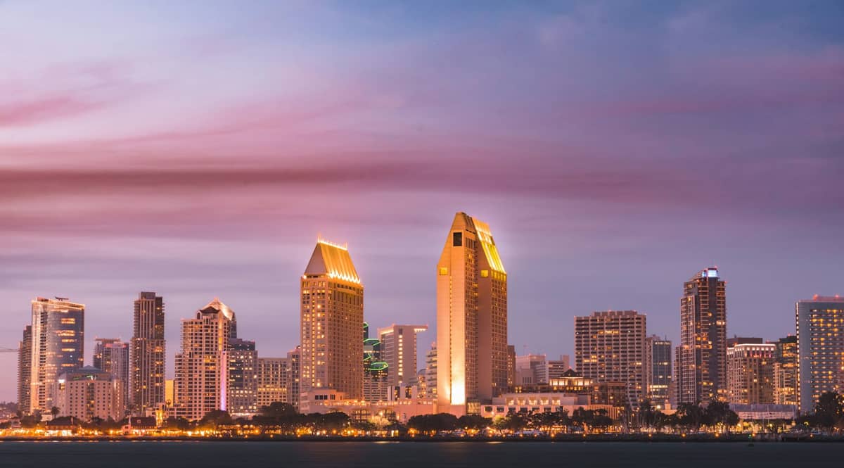 Weitläufige Ansicht der Skyline von San Diego im Sonnenuntergang mit der Uferpromenade im Vordergrund und einem Himmel in sanften Rosa- und Lilatönen.