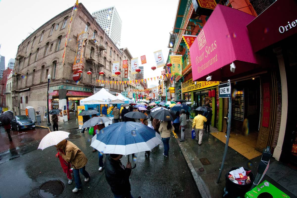 Menschenmassen laufen unter Regenschirmen an einem regnerischen Tag in San Franciscos Chinatown, während bunte Schilder und Laternen entlang der nassen Straßen zu sehen sind.
