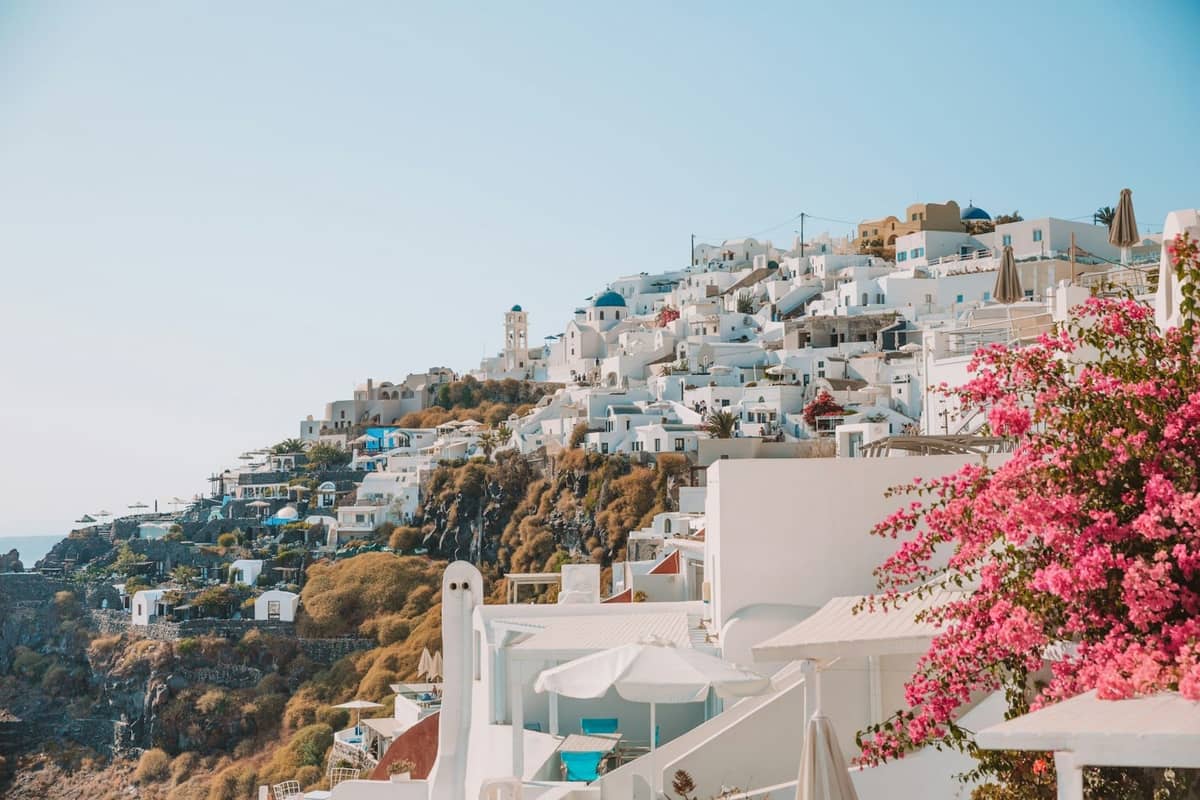 Klassischer Blick auf die Klippen von Santorin mit weiß getünchten Gebäuden, blauen Kuppeln und rosafarbenem Bougainvillea unter klarem blauem Himmel, mit einer ruhigen und angenehmen Atmosphäre.