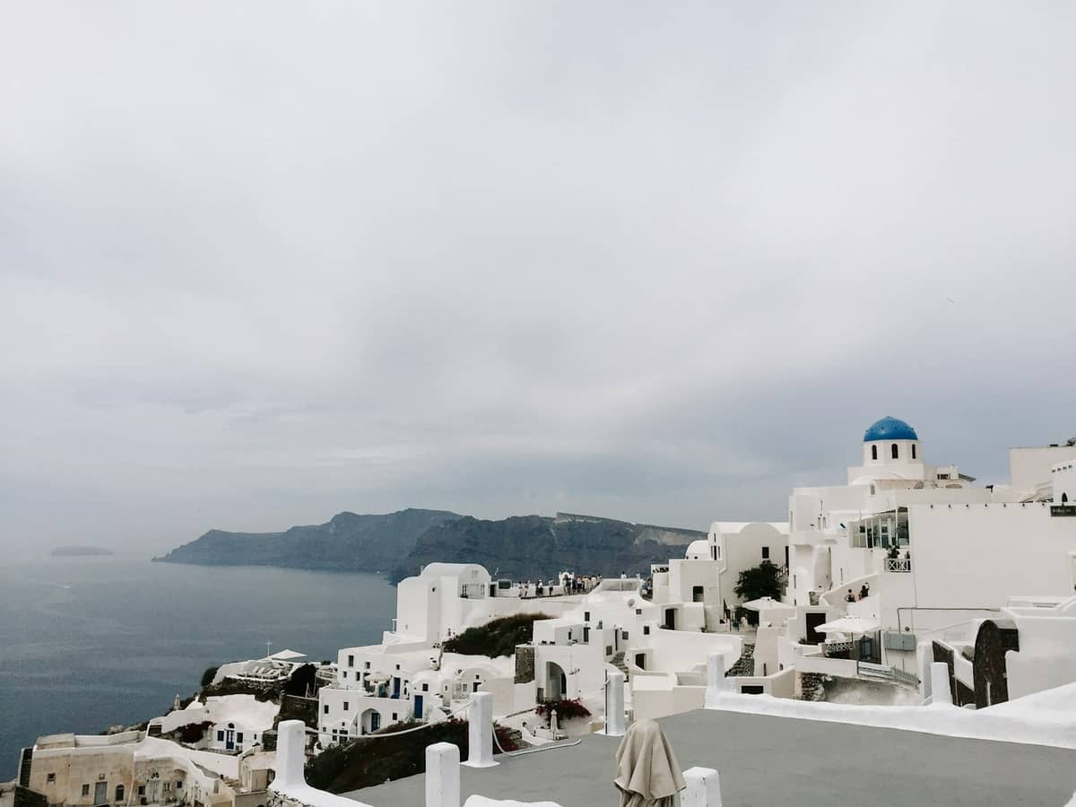 Ikonische weiß getünchte Gebäude mit blauen Kuppeln mit Blick auf das Meer und die Klippen in Santorini unter einem bewölkten Himmel.
