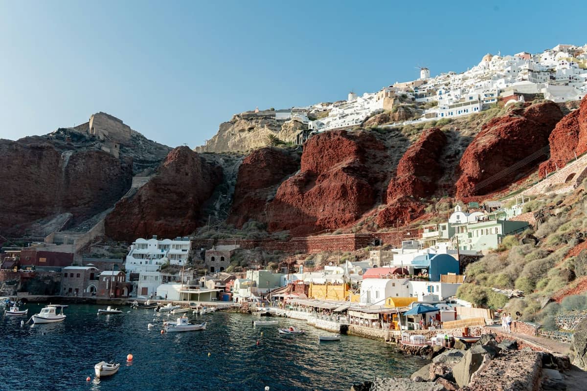 Hafen von Santorini mit traditionellen Restaurants am Wasser, Booten im Wasser und weiß getünchten Gebäuden am Hang, die saisonale Aktivitäten und lokale Unternehmen veranschaulichen.