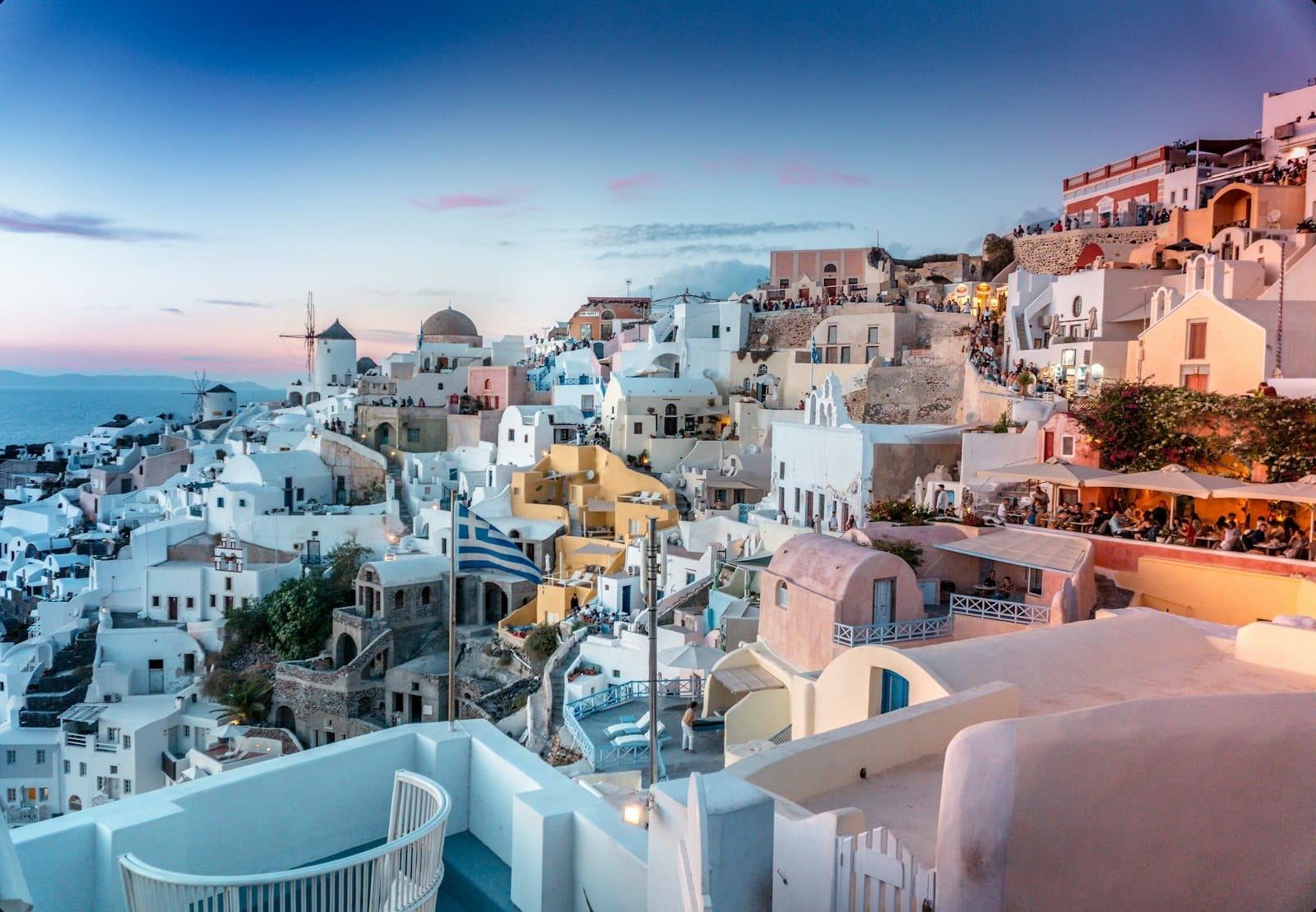 Vila de Santorini ao pôr do sol com céu em tons pastel, casinhas brancas, cúpulas azuis e o mar ao fundo, criando uma atmosfera sonhadora e convidativa.