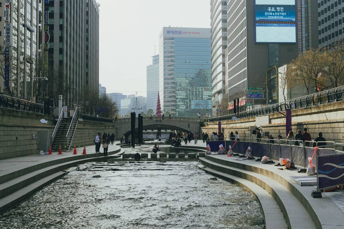 Córrego urbano Cheonggyecheon em Seul ladeado por altos edifícios de escritórios, pessoas caminhando e sentadas à beira da água em um dia nublado