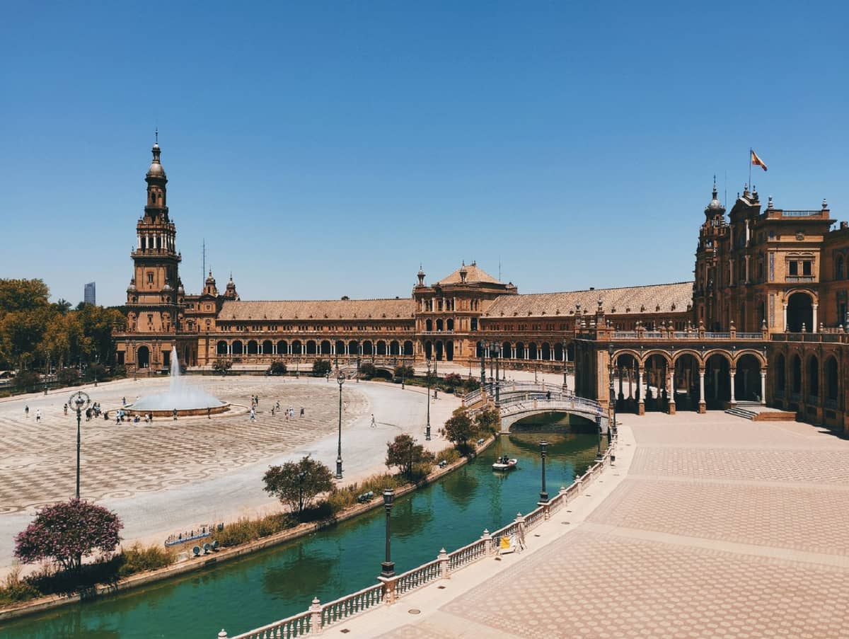 มุมกว้างของ Plaza de España แลนด์มาร์กเซบียา อาคารโค้ง สะพาน หอคอย คลองในวันแดดจัด