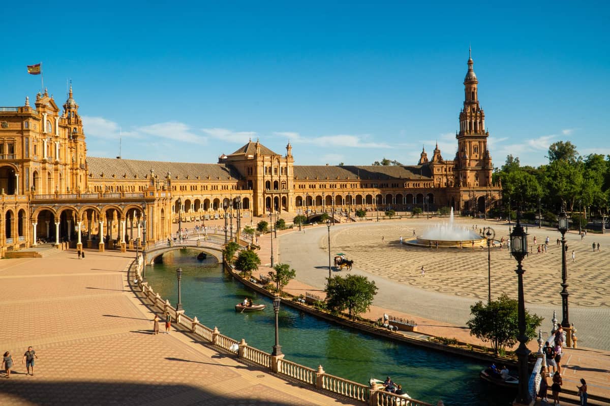 วิวกว้างของ Plaza de España ในเมืองเซบิยา ภายใต้ท้องฟ้าสีฟ้าใสในฤดูร้อน พร้อมแสงแดดจ้า ลานกว้างโล่งและแห้ง และผู้คนที่เดินอยู่รอบๆ