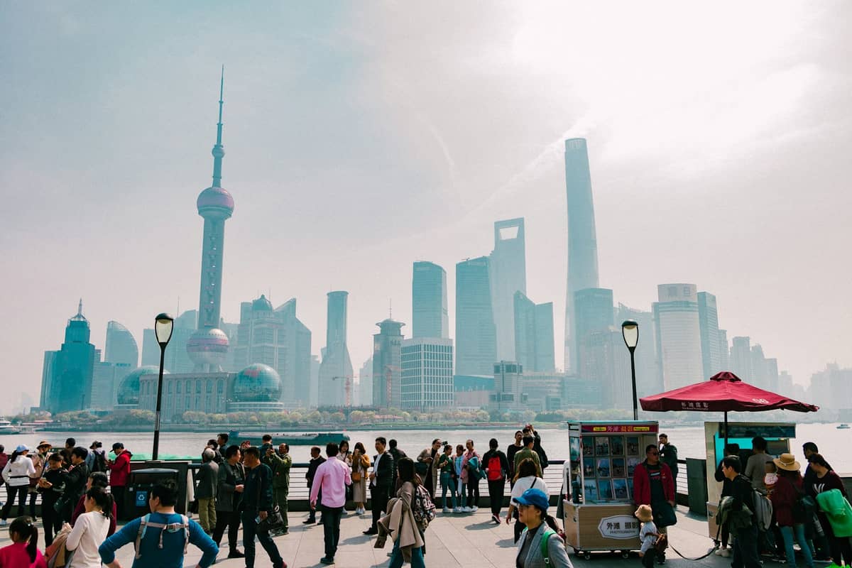 Eine belebte Uferpromenade in Shanghai mit vielen Spaziergängern und den modernen Wolkenkratzern Shanghais, die sich im sommerlichen Dunst als Silhouette abzeichnen.