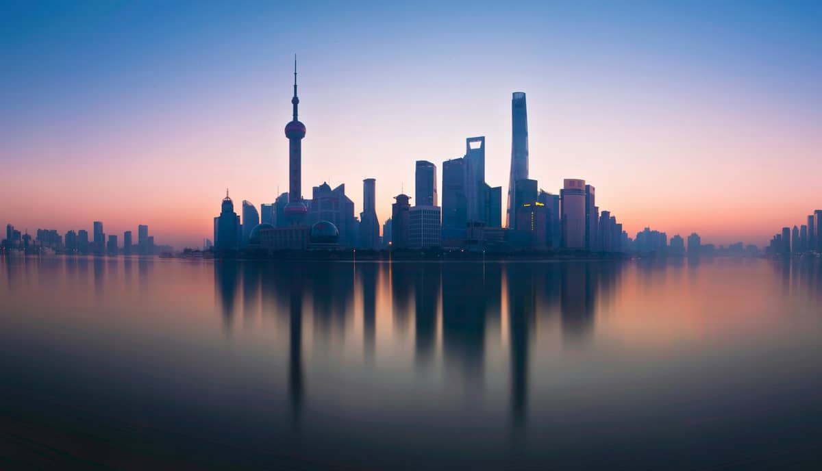 Weiter Blick auf die Skyline Shanghais bei Sonnenaufgang mit dem Oriental Pearl Tower und modernen Wolkenkratzern, die sich im ruhigen Huangpu-Fluss spiegeln.