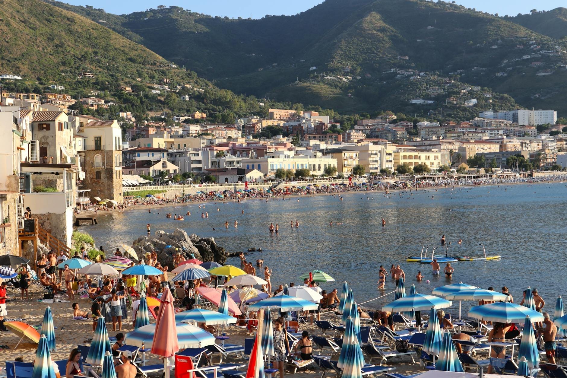 Überfüllter sizilianischer Strand im Sommer mit Sonnenschirmen, Sonnenbadenden, Schwimmern im Meer sowie Hügeln und Ortschaft im Hintergrund.