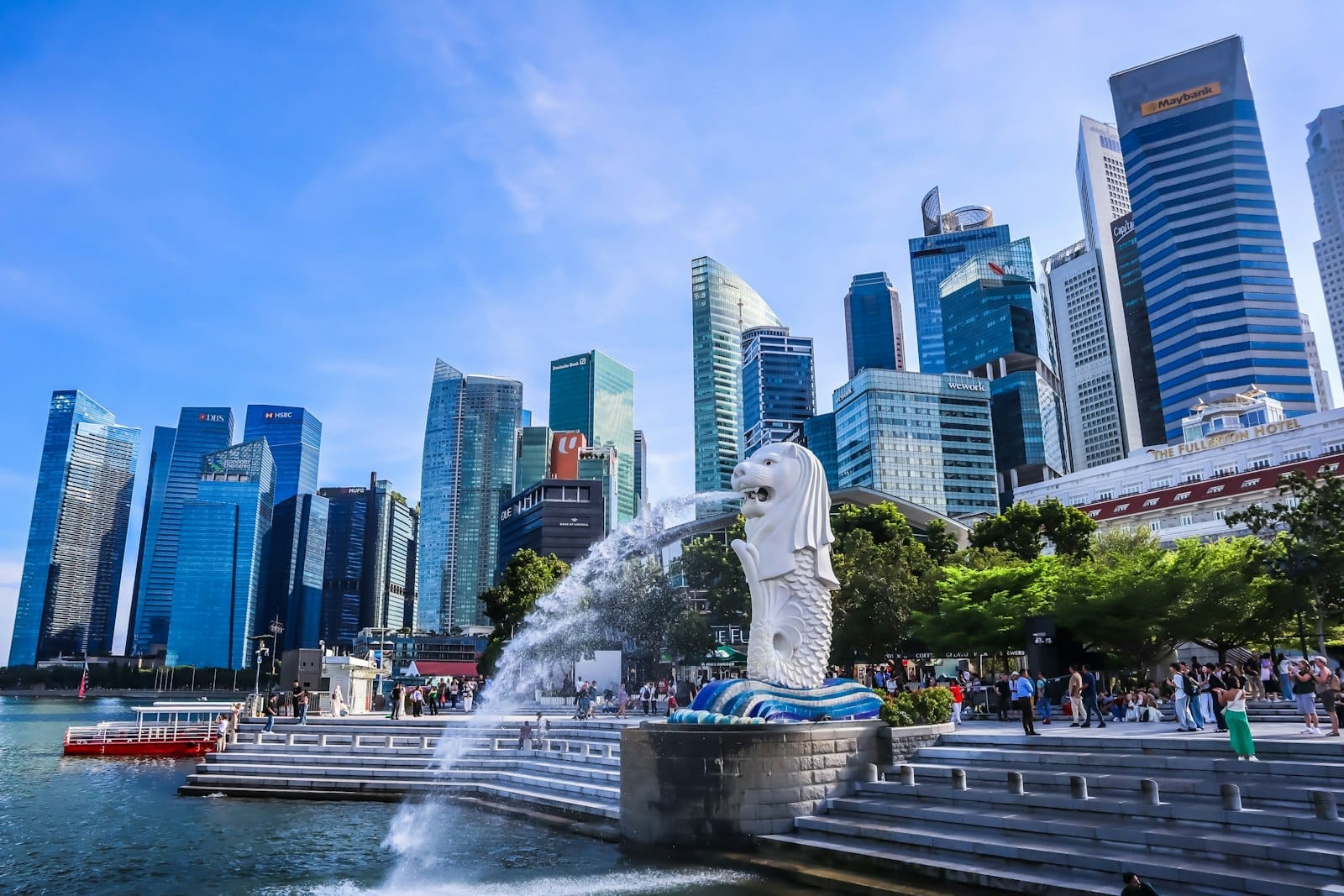 Multidão de turistas ao lado da famosa estátua Merlion com o skyline moderno de Singapura e céu azul ao fundo, mostrando a atmosfera vibrante da cidade.