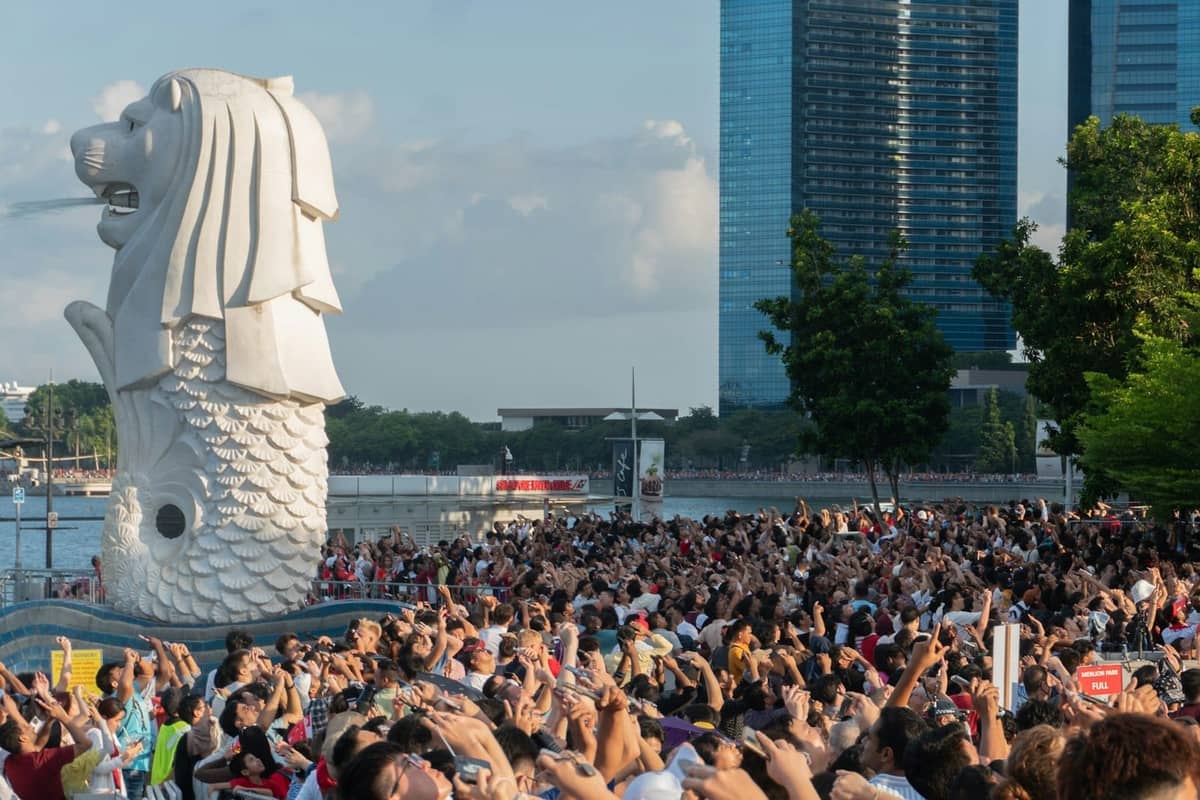 Large crowd of tourists taking photos at Singapore's famous Merlion Park, with the Merlion statue and modern skyscrapers in the background on a sunny day.