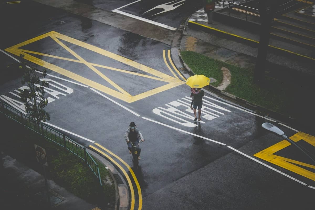 Pedestrians and a cyclist navigate a wet Singapore street, yellow road markings and an umbrella visible after recent rainfall.