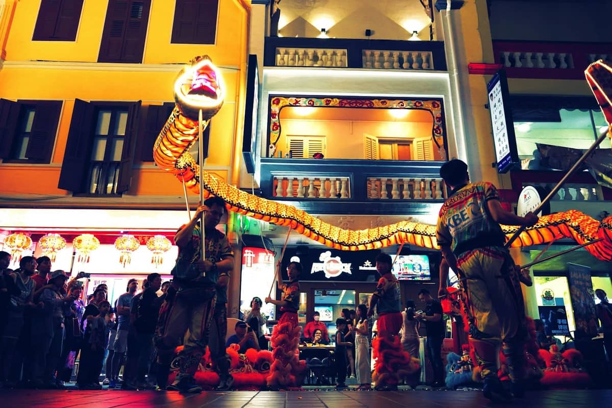 Performers in traditional costume hold a glowing dragon during a night parade in Singapore's Chinatown, with colorful shophouses and lanterns in the background.