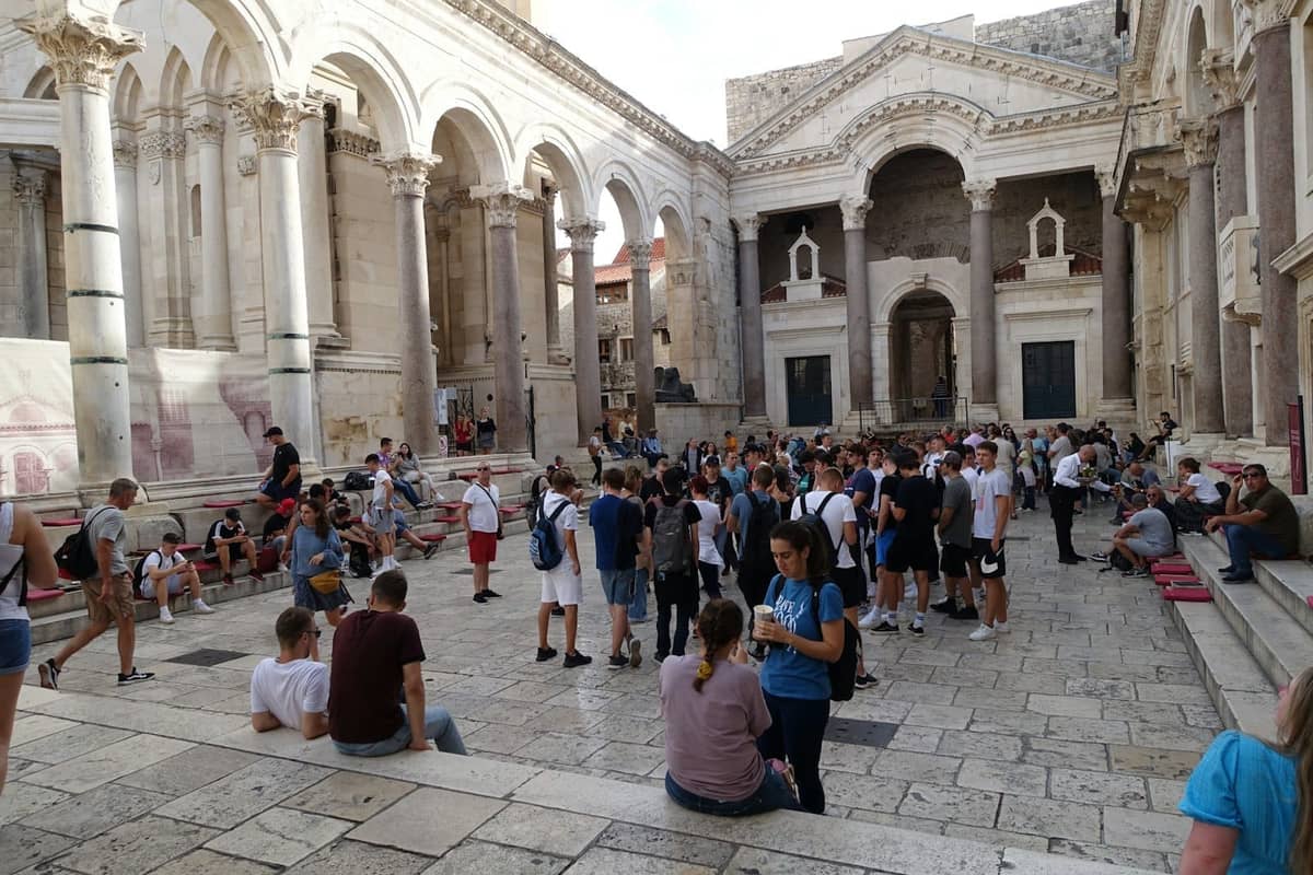 Eine belebte Sommermenge füllt den Peristyl-Hof des Diokletianpalastes in Split, Kroatien, mit Menschen, die zwischen der antiken Marmorarchitektur sitzen und stehen.