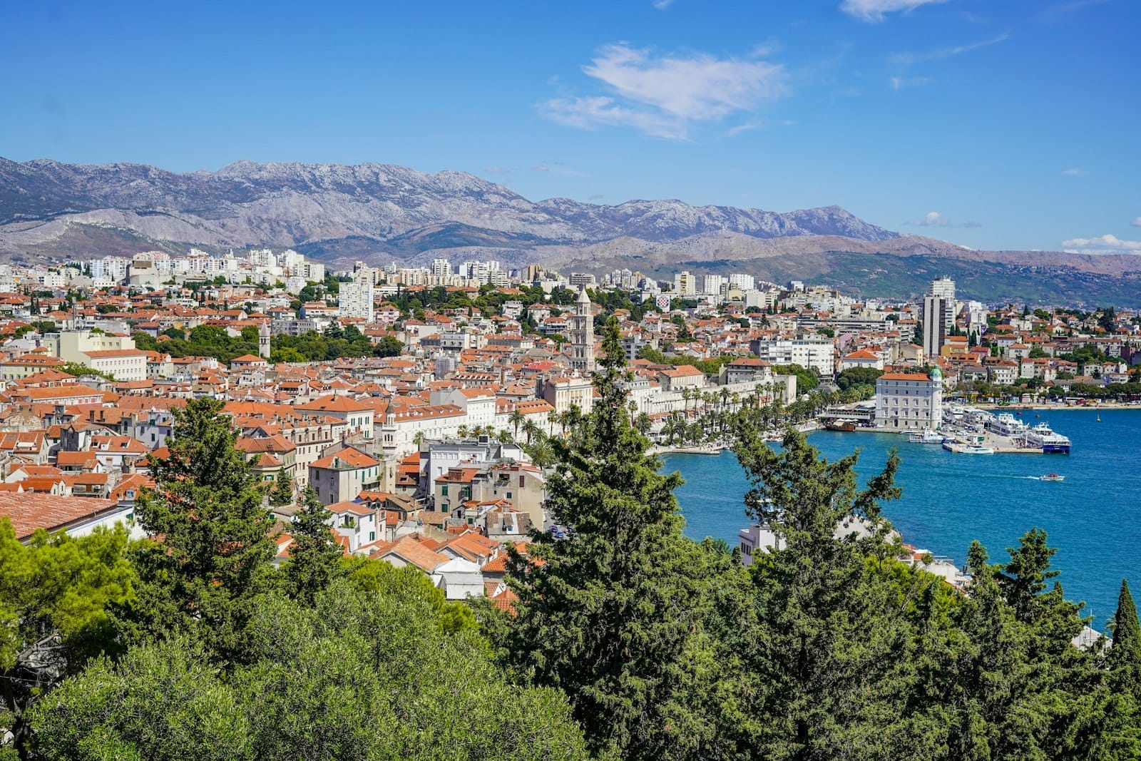 Vista panorâmica de Split, na Croácia, com o centro histórico, o porto ladeado de palmeiras, o Mar Adriático azul e montanhas ao fundo sob um céu azul intenso.