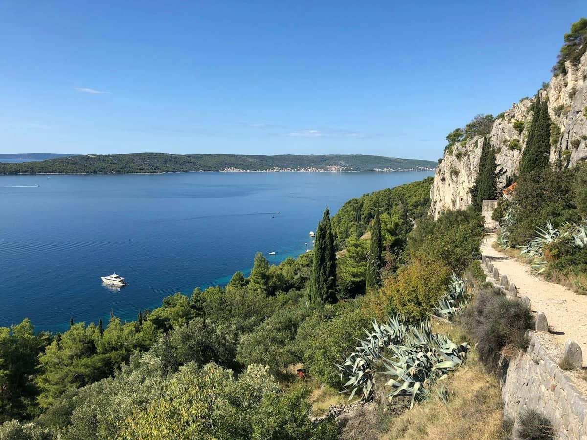 Malerischer Küstenweg in Split mit klarem blauem Wasser, üppigem Grün und felsigen Klippen unter einem sonnigen Himmel.