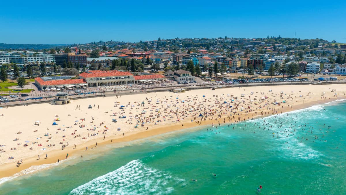 Vista aérea de Bondi Beach en un soleado día de verano lleno de bañistas, agua turquesa cristalina y los suburbios del este de Sídney al fondo.