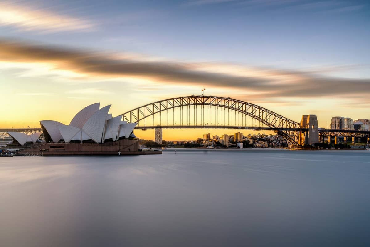 La Ópera de Sídney y el Harbour Bridge al amanecer con agua tranquila y el horizonte de la ciudad al fondo.