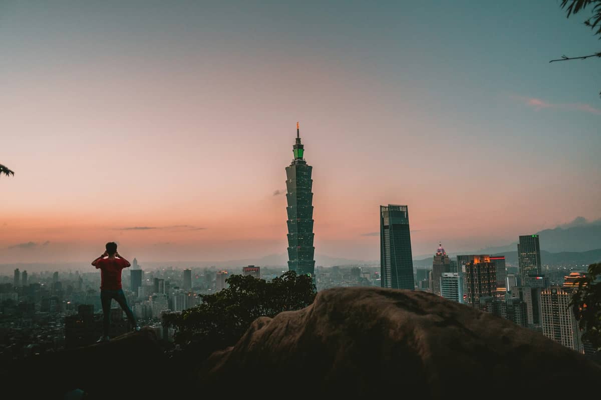 Skyline von Taipeh bei Sonnenuntergang mit dem Taipei 101 im Mittelpunkt, fotografiert vom Elephant Mountain mit einer Person, die die Aussicht genießt.