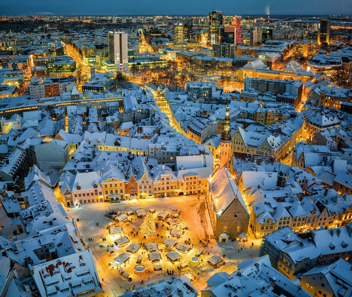 Luftaufnahme der schneebedeckten Tallinner Altstadt in der Abenddämmerung, mit festlichen Lichtern, die den Rathausplatz und den zentralen Weihnachtsmarkt erleuchten.
