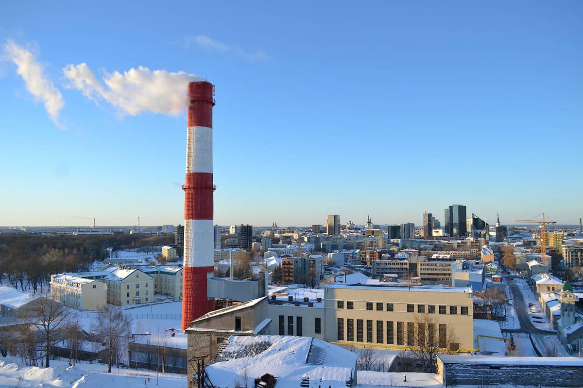 Vista panorámica invernal de Tallinn con una chimenea roja y blanca emitiendo vapor, tejados cubiertos de nieve y un cielo azul despejado.
