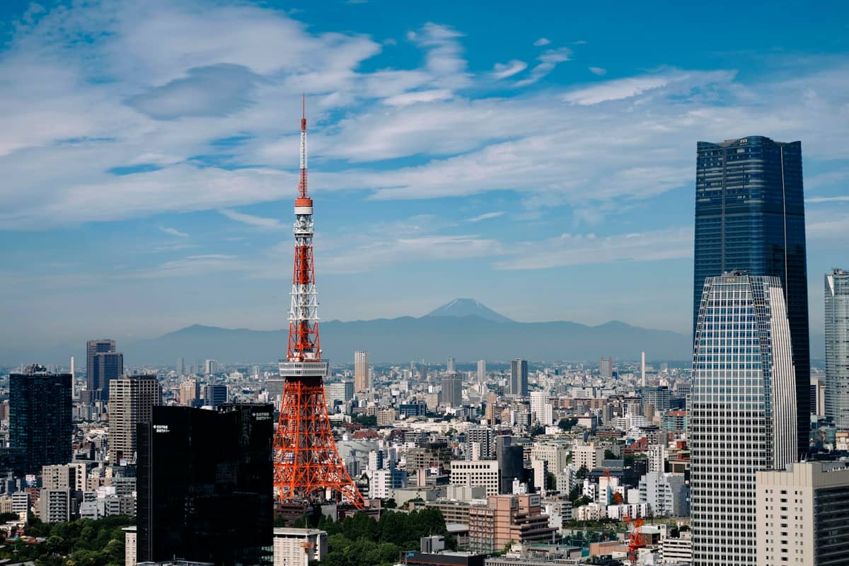 Daytime view of Tokyo skyline with Tokyo Tower and city buildings under partly cloudy sky, Mount Fuji visible in the distance.