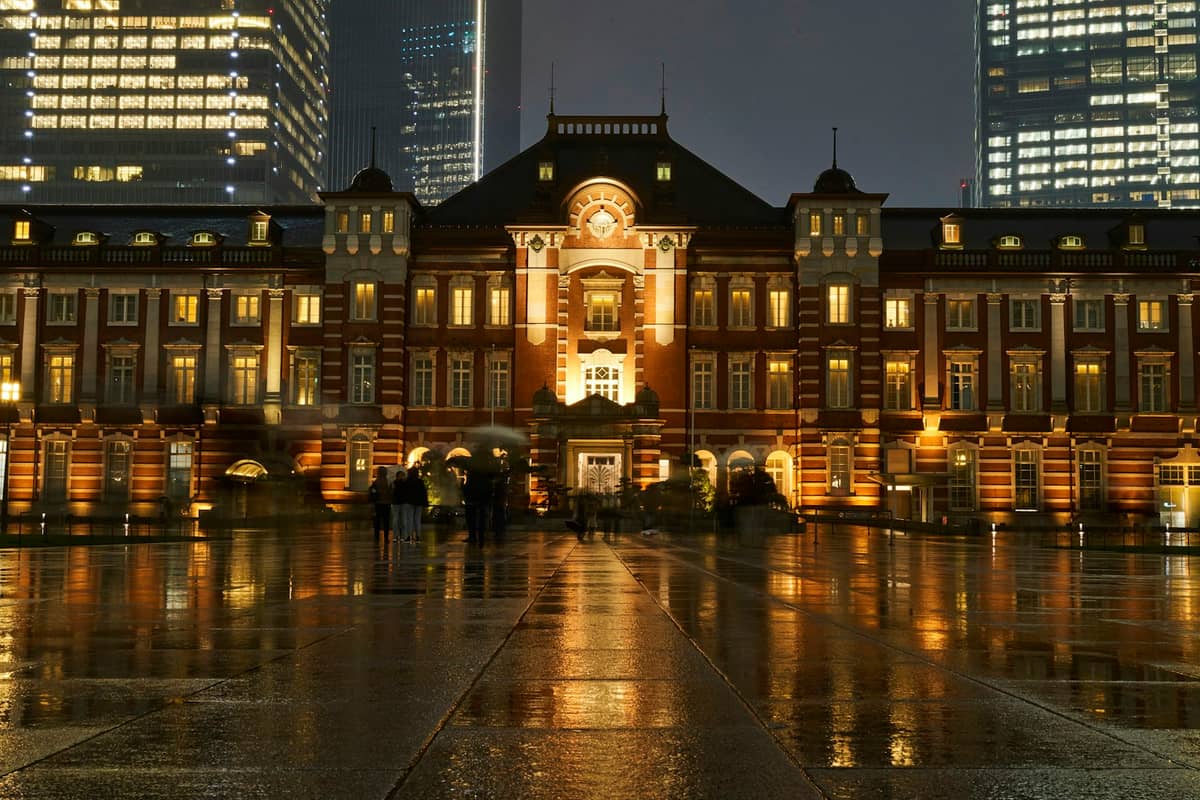 Tokyo Station at night, warmly lit with reflections on wet ground, surrounded by modern skyscrapers and sparse people, evoking a crisp winter evening.