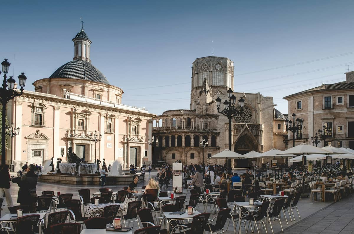 Tavolini di un caffè all'aperto in Plaza de la Virgen con la Cattedrale di Valencia e la fontana sullo sfondo, sotto un cielo terso che trasmette tutta la vivacità della città.