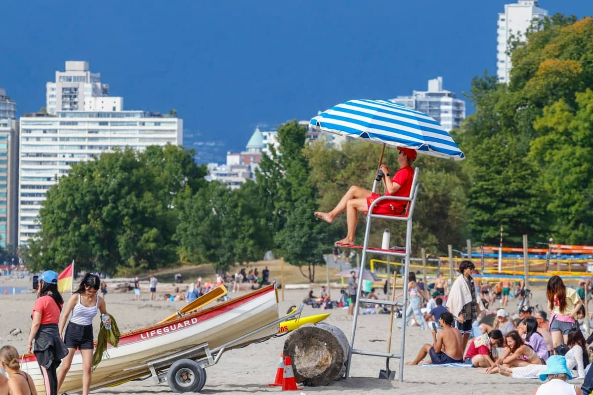 Überfüllter Strand in Vancouver mit Rettungsschwimmer, Sonnenbadenden und Stadtgebäuden im Sommer bei klarem Wetter.